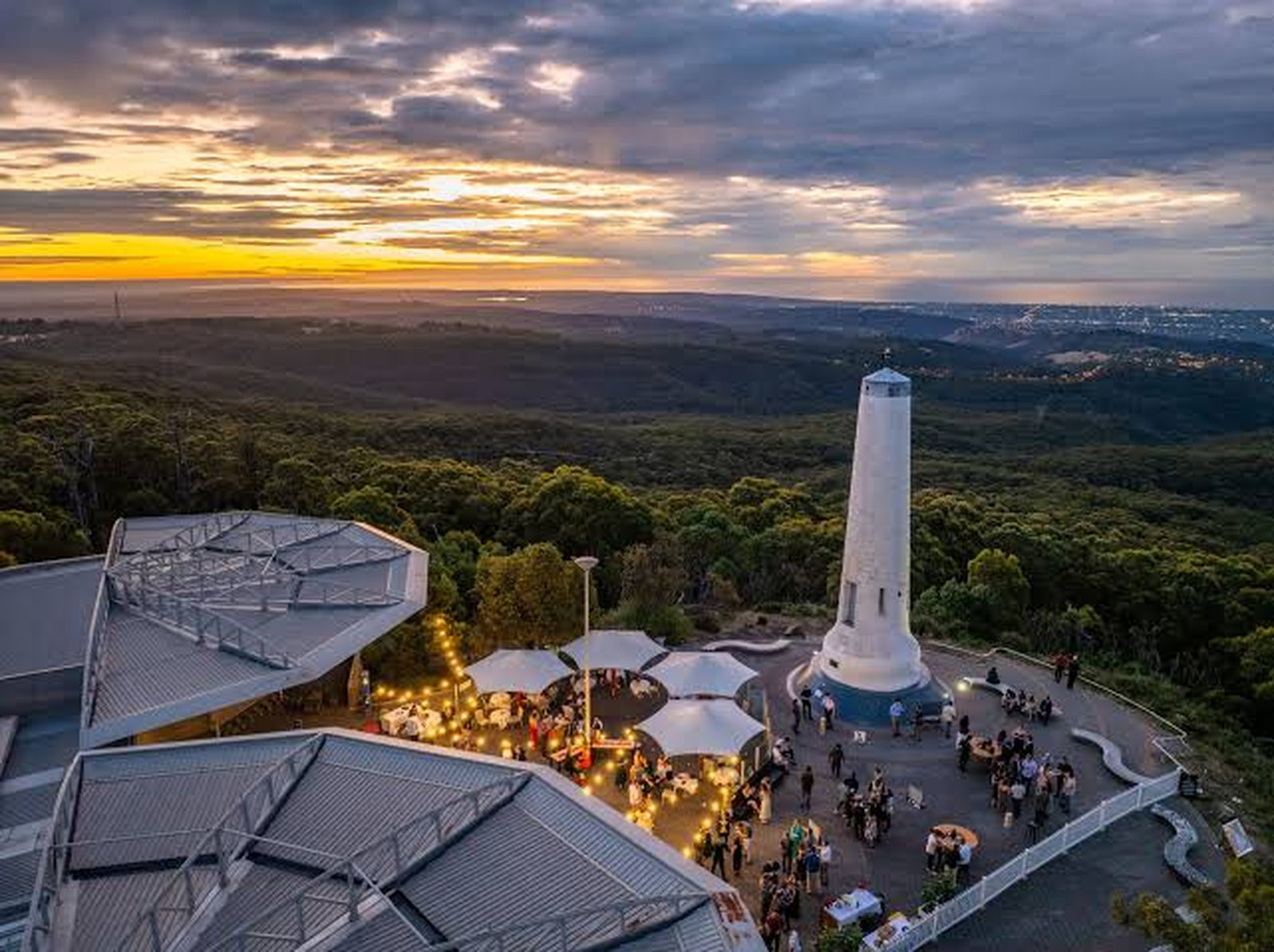 A hilltop plaza and white monument at dusk, looking out over forested hills toward a city