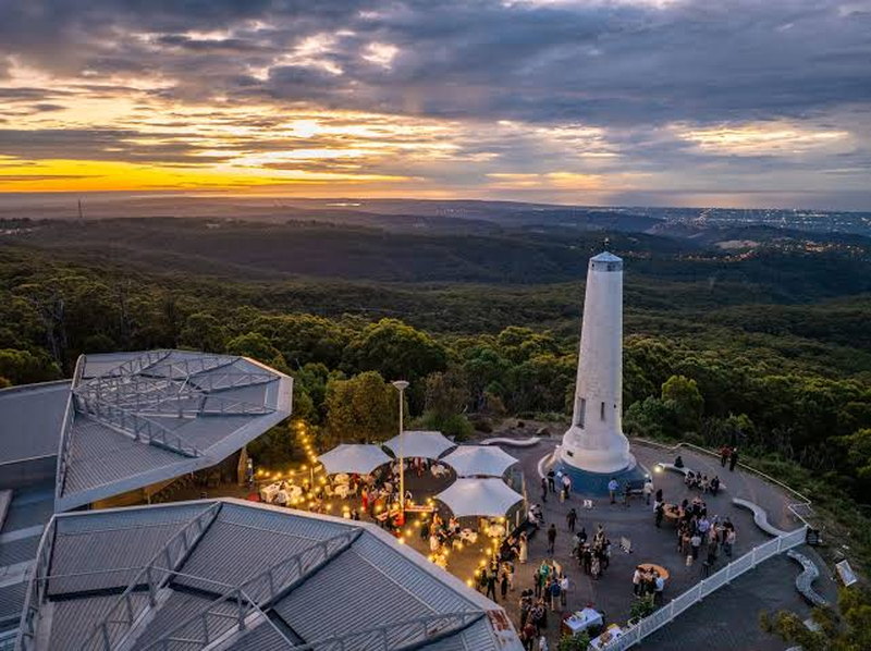 A hilltop plaza and white monument at dusk, looking out over forested hills toward a city